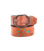 The Upswing II by Bed|Stü, a handcrafted leather belt featuring decorative cutouts that reveal turquoise accents and finished with a hexagonal metal buckle, is coiled on a white background.