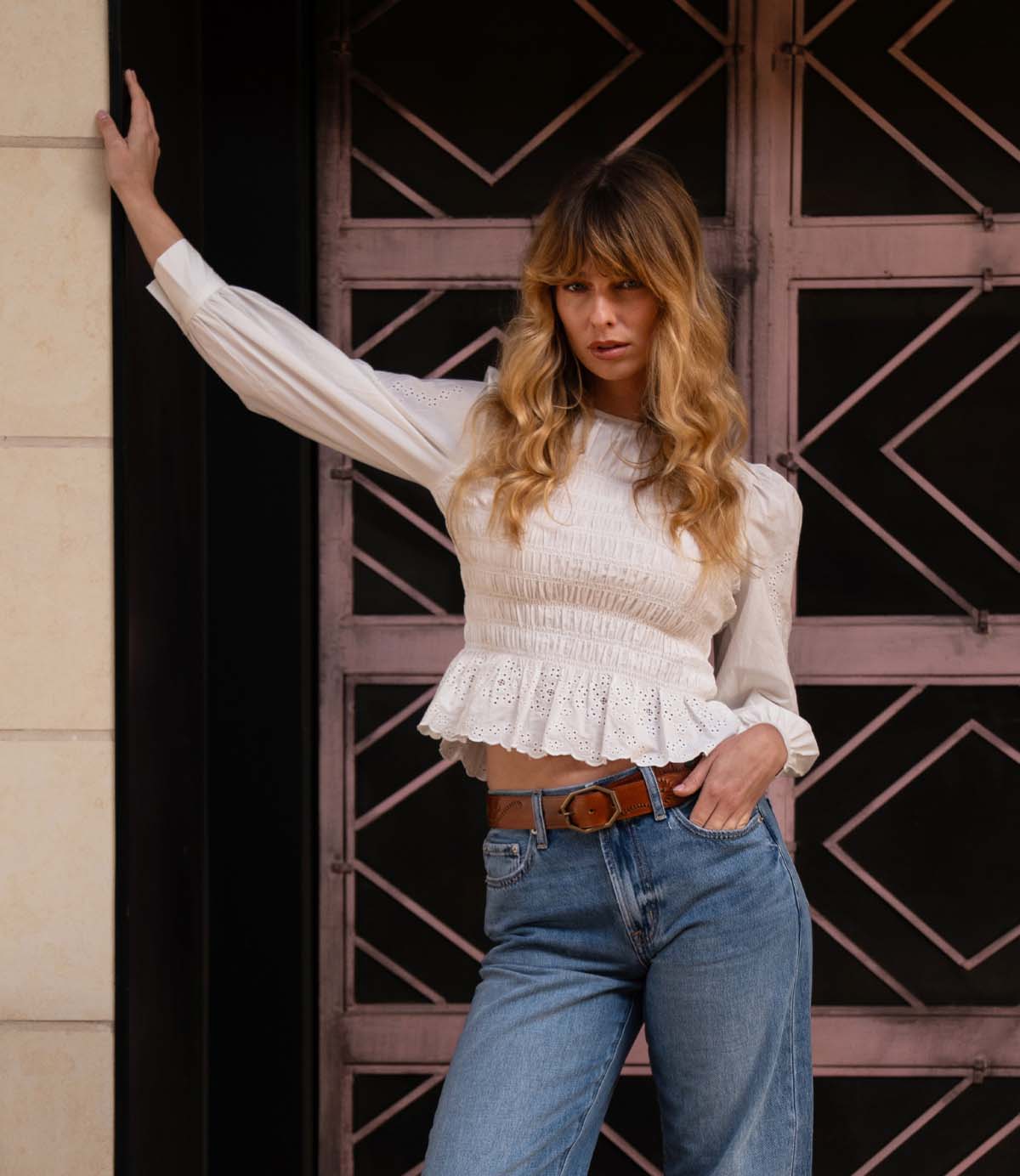 A woman with long blonde hair poses in front of a patterned metal door, wearing a white blouse and blue jeans paired with the Bed|Stü Upswing II artisanal belt, one hand on her hip and the other resting on the wall.