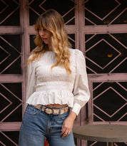 A woman with wavy blonde hair, dressed in a white smocked blouse and blue jeans, stands near a round table wearing the Bed|Stü Upswing II artisanal belt, with a pink window frame featuring diamond patterns in the background.