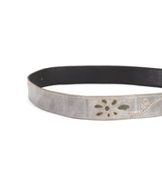 The Upswing II by Bed|Stü is a silver leather belt featuring decorative cut-out floral patterns with green accents, shown partially coiled on a white background.