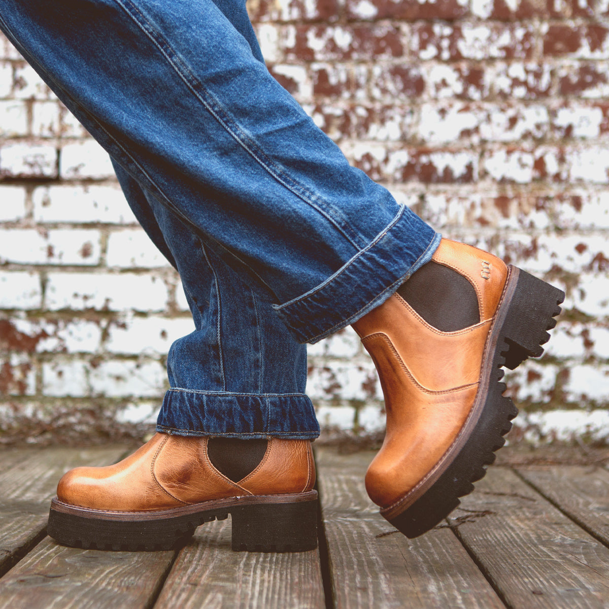 A person wearing cuffed blue jeans and Bed|Stü Valda Hi tan leather Chelsea boots with a comfortable platform sole stands on a wooden deck in front of a white brick wall.