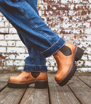 A person wearing cuffed blue jeans and Bed|Stü Valda Hi tan leather Chelsea boots with a comfortable platform sole stands on a wooden deck in front of a white brick wall.