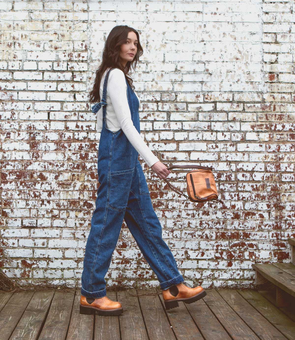 A woman wearing blue denim overalls, a white long-sleeve shirt, and the Bed|Stü Valda Hi comfortable brown Chelsea boots with a platform sole stands on a wooden deck in front of a weathered white brick wall, holding a small tan bag.