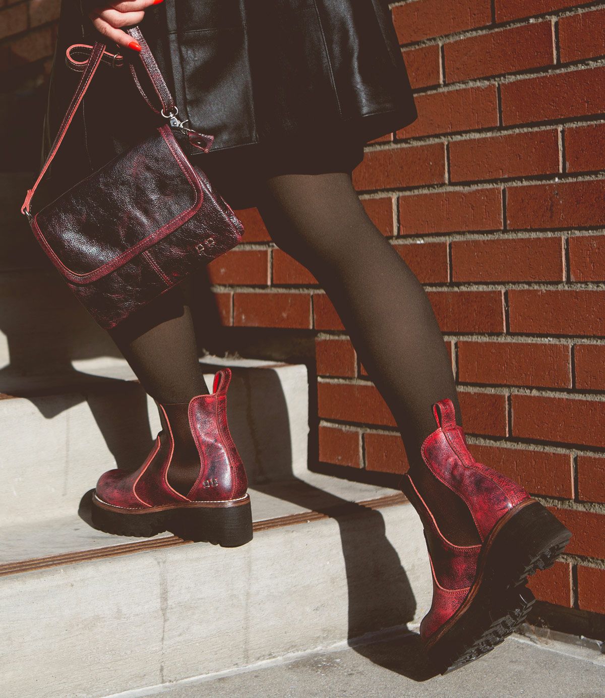 A person wearing black tights, a black skirt, and comfortable Bed|Stü Valda Hi red leather boots with a platform sole, while carrying a matching red handbag, walks up concrete steps beside a brick wall.