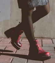 A person wearing black tights, a dark skirt, and comfortable Bed|Stü Valda Hi red leather Chelsea boots with a chunky platform sole walks on a brick sidewalk next to a concrete wall.