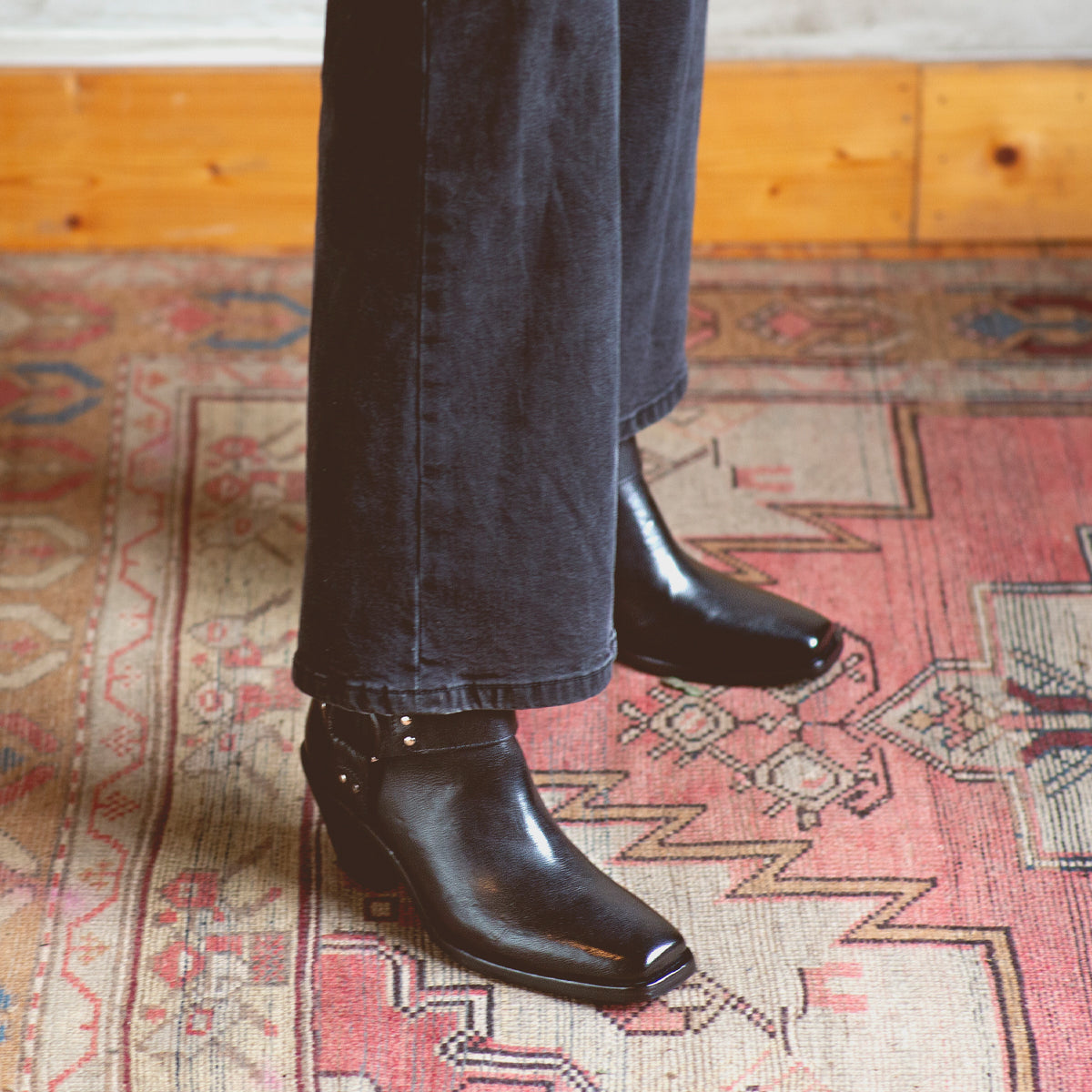 Wearing Bed|Stü's Vamoose sleek black square-toe boots and black pants, the person stands on a patterned rug with a wooden baseboard in the background.