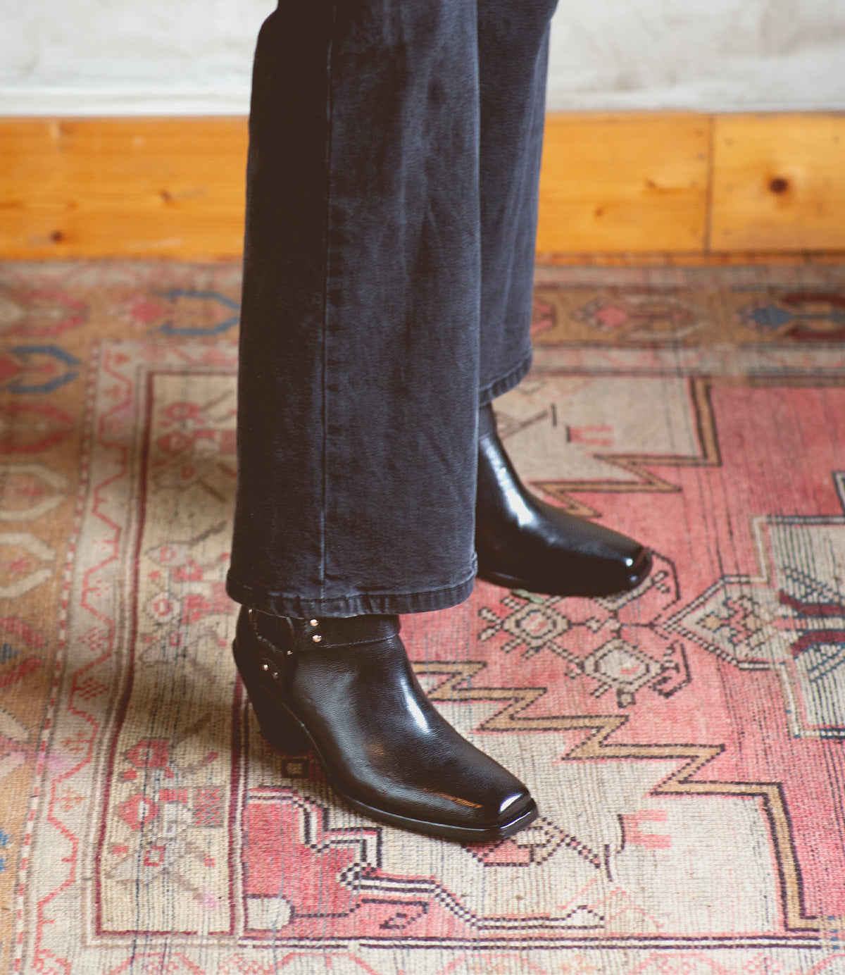 Wearing Bed|Stü's Vamoose sleek black square-toe boots and black pants, the person stands on a patterned rug with a wooden baseboard in the background.