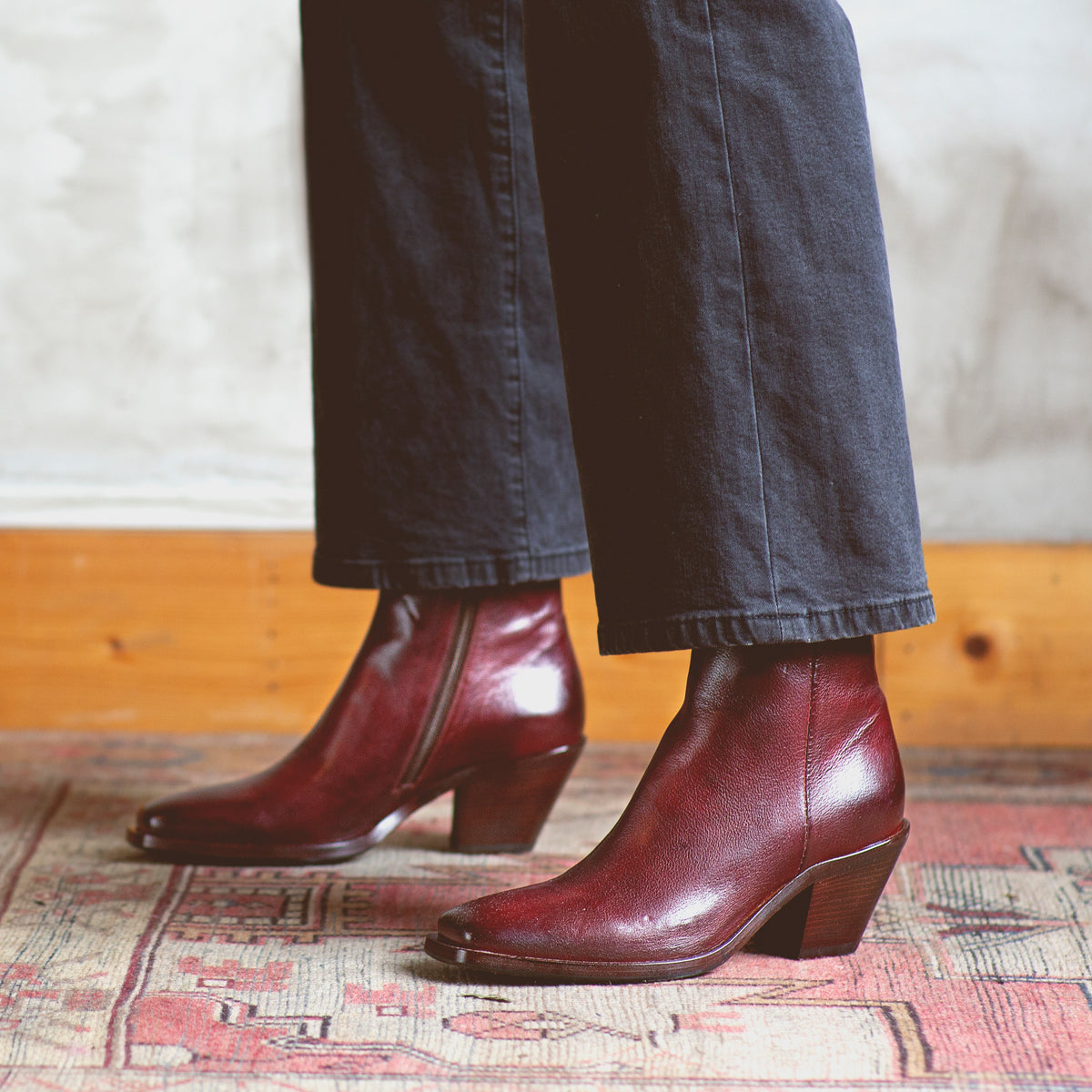 A person wearing dark pants and Bed|Stü Vendue burgundy square-toe heeled ankle boots stands on a patterned rug, with a wooden baseboard in the background.