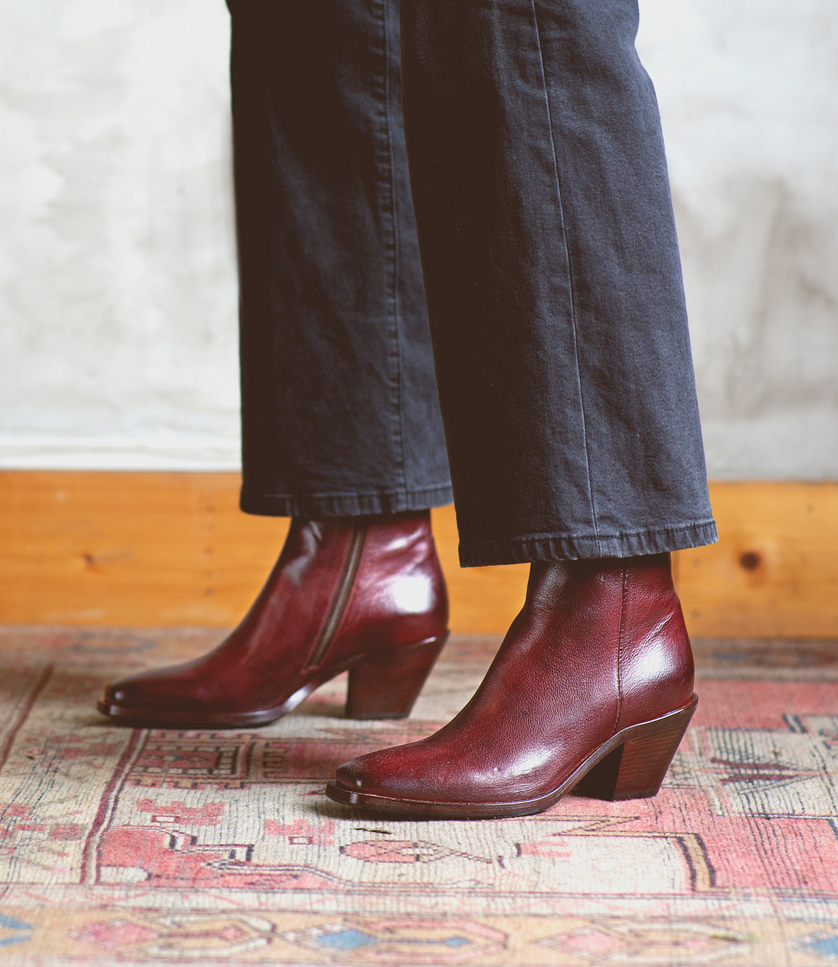 A person wearing dark pants and Bed|Stü Vendue burgundy square-toe heeled ankle boots stands on a patterned rug, with a wooden baseboard in the background.