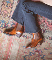 A person wearing Bed|Stü Vendue brown heeled Chelsea boots with a square toe and dark pants sits cross-legged on a patterned rug with geometric designs.