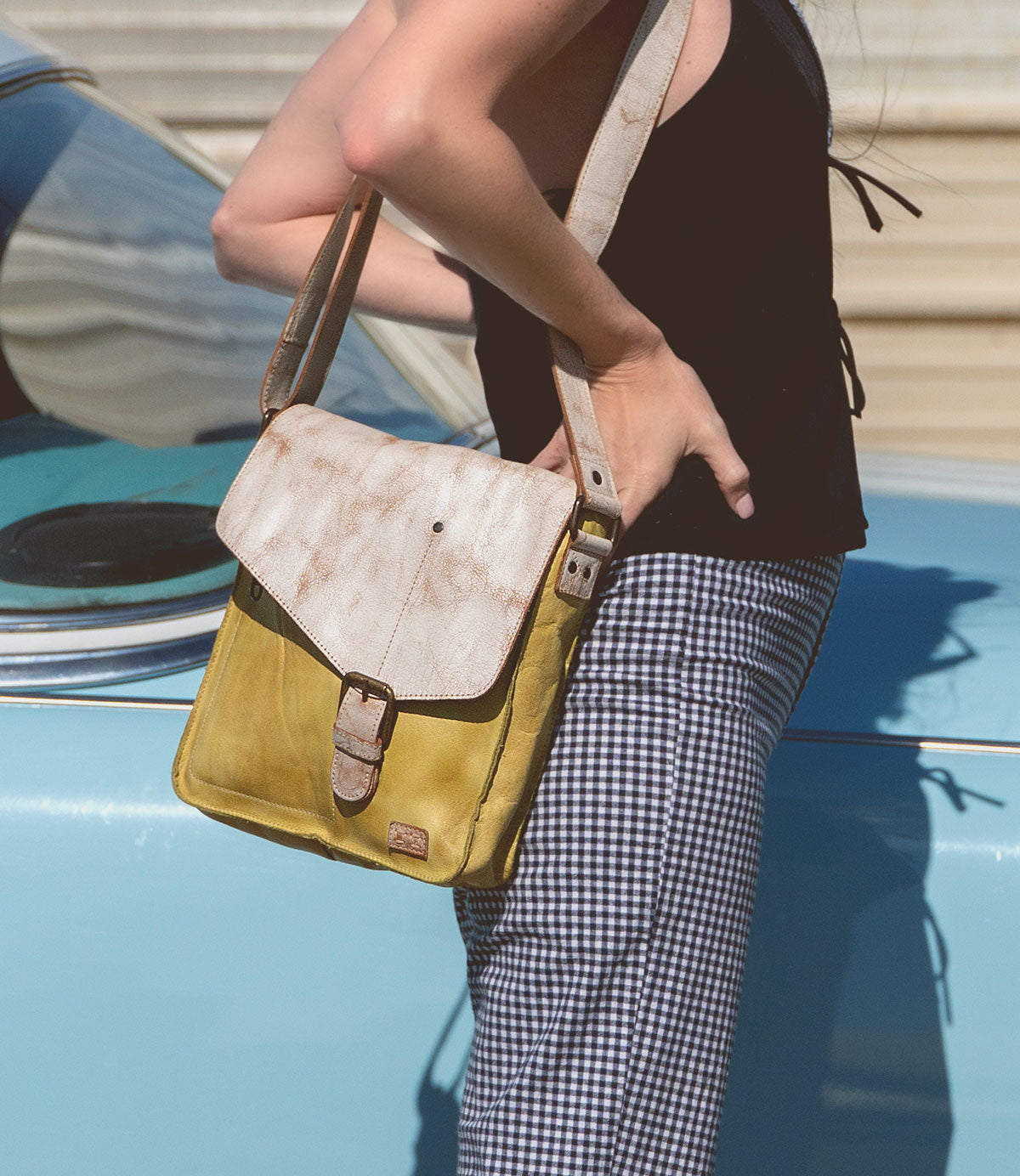 A person stands beside a light blue car, dressed in a black top and checkered pants, carrying the Bed|Stü Venice Beach yellow and beige crossbody bag with an adjustable strap.