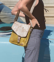 A person stands beside a light blue car, dressed in a black top and checkered pants, carrying the Bed|Stü Venice Beach yellow and beige crossbody bag with an adjustable strap.