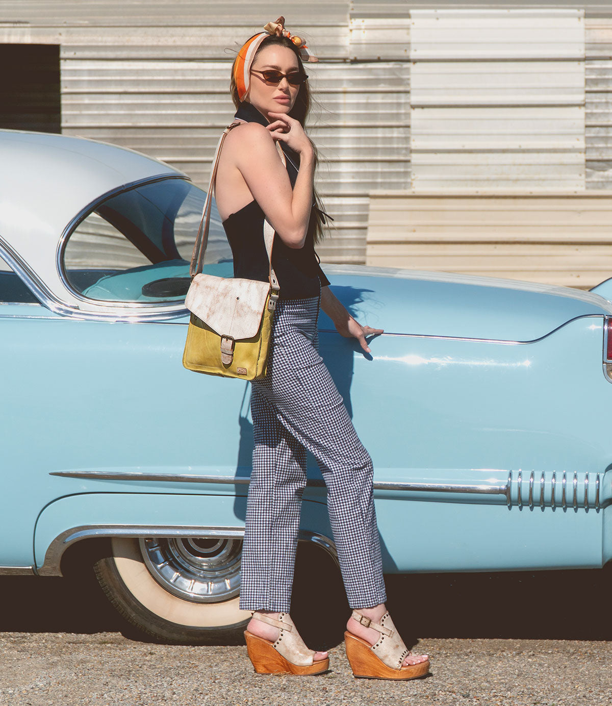 A woman stands beside a vintage blue car, wearing sunglasses, a headscarf, black top, checkered pants, wedge sandals, and the Bed|Stü Venice Beach full-grain leather crossbody bag with an adjustable strap.
