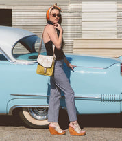 A woman stands beside a vintage blue car, wearing sunglasses, a headscarf, black top, checkered pants, wedge sandals, and the Bed|Stü Venice Beach full-grain leather crossbody bag with an adjustable strap.