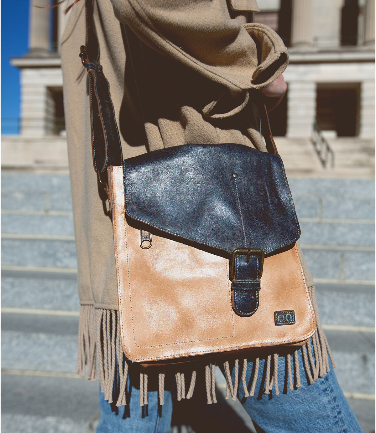 A person wearing a tan fringed coat carries the Bed|Stü Venice Beach satchel-style crossbody bag in tan and black full-grain leather, featuring an adjustable strap with buckle, while standing on outdoor stone steps.