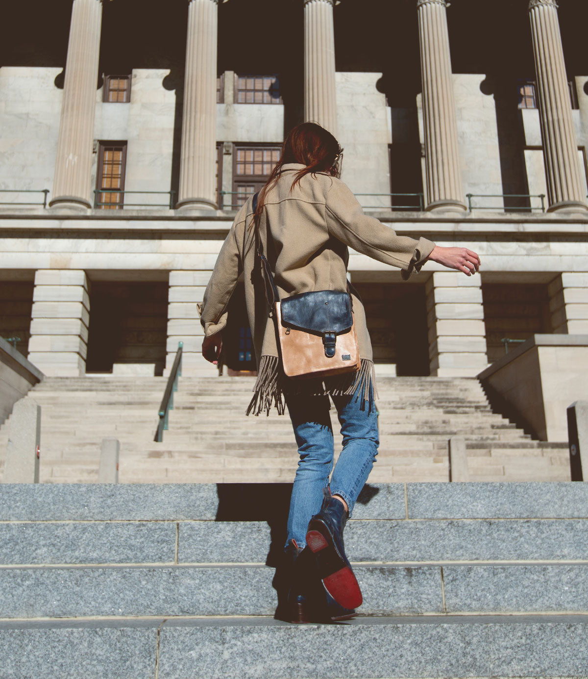 A person wearing a tan coat and blue jeans walks up stone steps toward a building with large columns, carrying the Bed|Stü Venice Beach crossbody bag crafted from stylish full-grain leather with an adjustable strap.