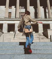 A person wearing a tan coat and blue jeans walks up stone steps toward a building with large columns, carrying the Bed|Stü Venice Beach crossbody bag crafted from stylish full-grain leather with an adjustable strap.