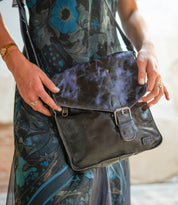 A person in a floral dress holds the Bed|Stü Venice Beach crossbody bag, crafted from black full-grain leather with an adjustable strap, flap closure, buckle accent, and stitched details.