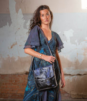 A woman stands indoors in front of a cracked, faded wall, wearing a blue patterned dress and carrying the Bed|Stü Venice Beach crossbody bag—crafted from full-grain leather with an adjustable strap—her expression neutral as she poses.