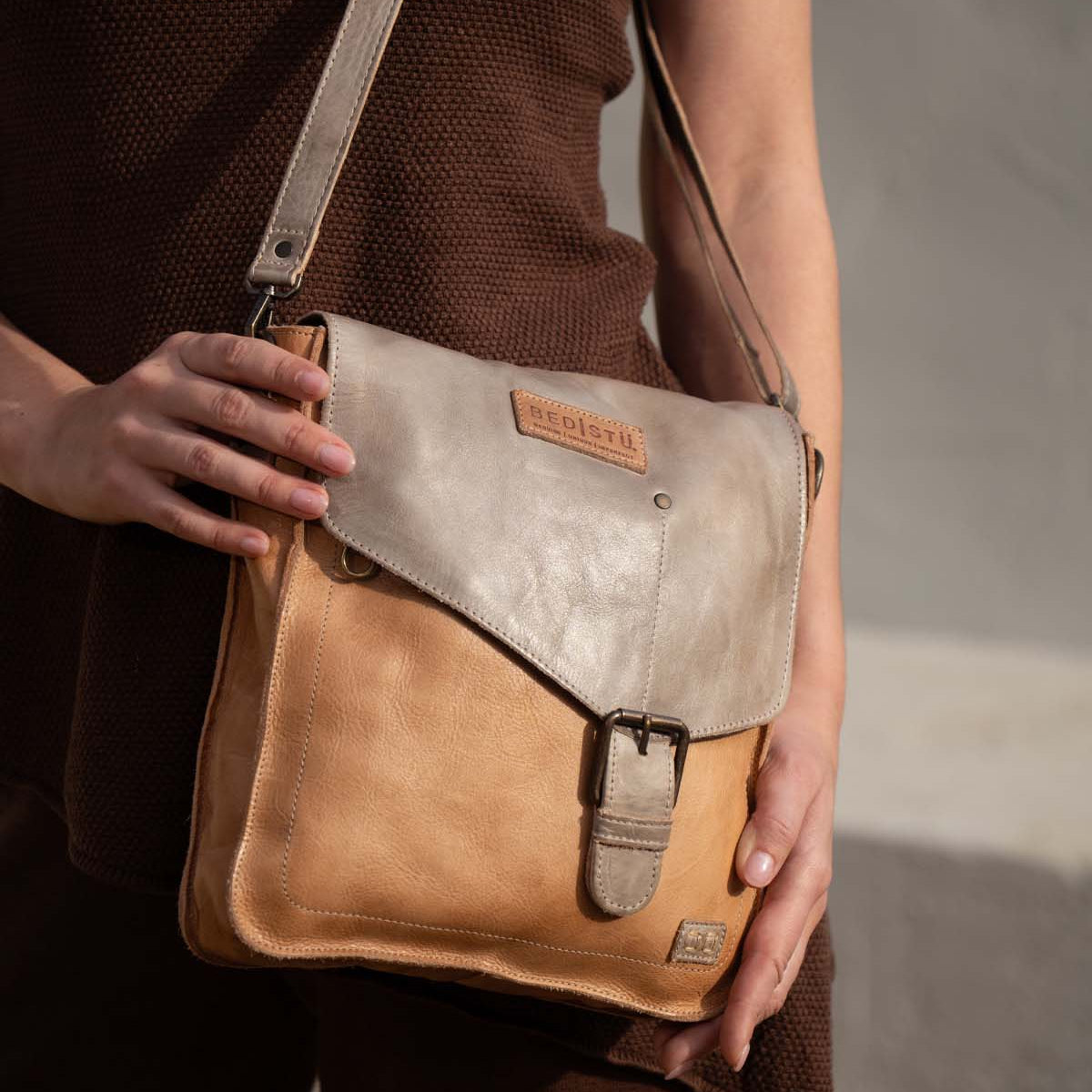 A person holds the Bed|Stü Venice Beach, a two-tone full-grain leather crossbody bag featuring an adjustable strap, buckle clasp, and embossed logo, while standing against a neutral background.