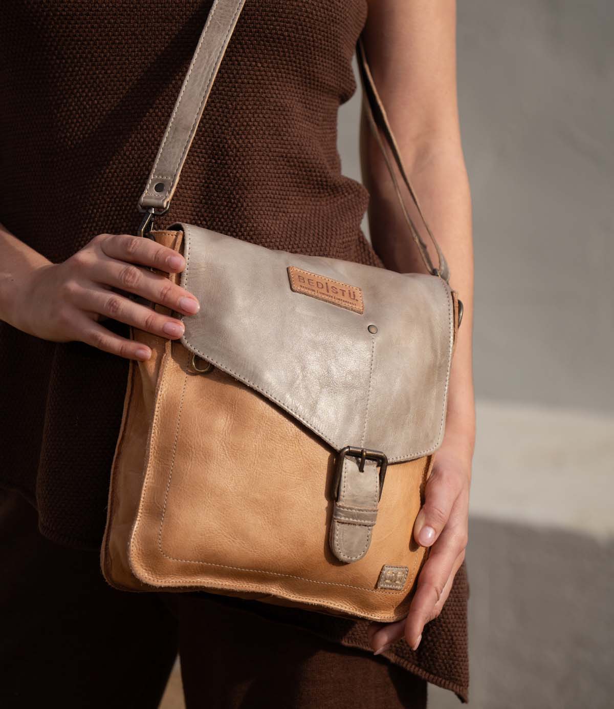 A person holds the Bed|Stü Venice Beach, a two-tone full-grain leather crossbody bag featuring an adjustable strap, buckle clasp, and embossed logo, while standing against a neutral background.