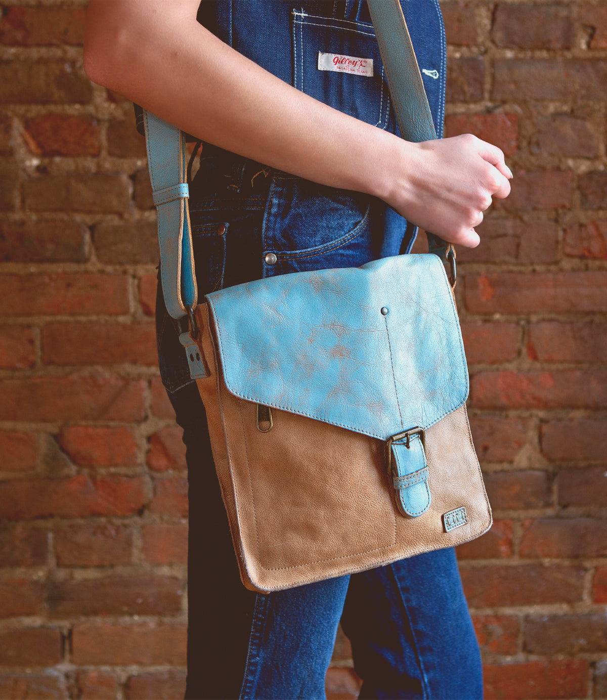 A person wearing denim overalls holds the Bed|Stü Venice Beach shoulder bag—crafted from tan and light blue full-grain leather with an adjustable strap—against a brick wall background.