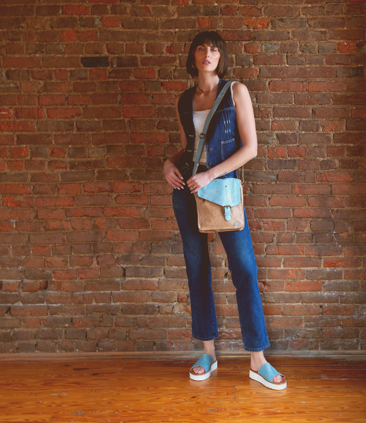 A woman in blue denim overalls, a white top, and blue sandals stands against a brick wall, holding the Bed|Stü Venice Beach crossbody bag in tan and blue with an adjustable strap.