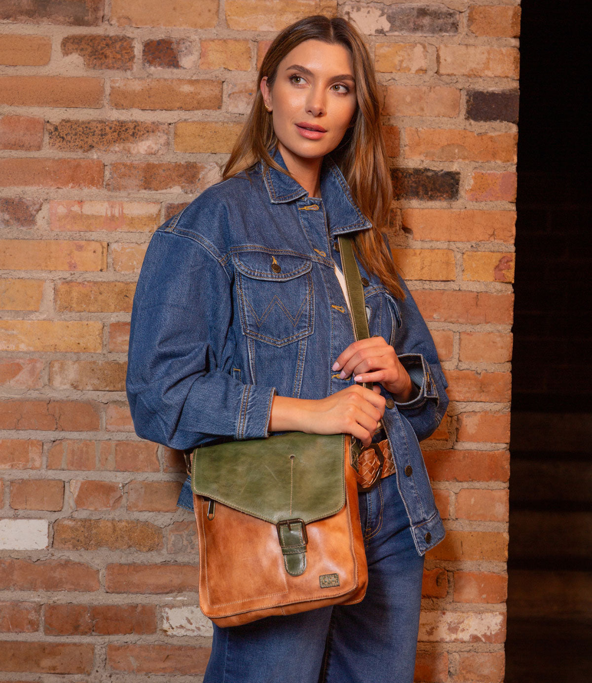 A woman stands by a brick wall, wearing a blue denim jacket and jeans, and carries the Bed|Stü Venice Beach crossbody bag in brown and green full-grain leather with an adjustable strap across her body.