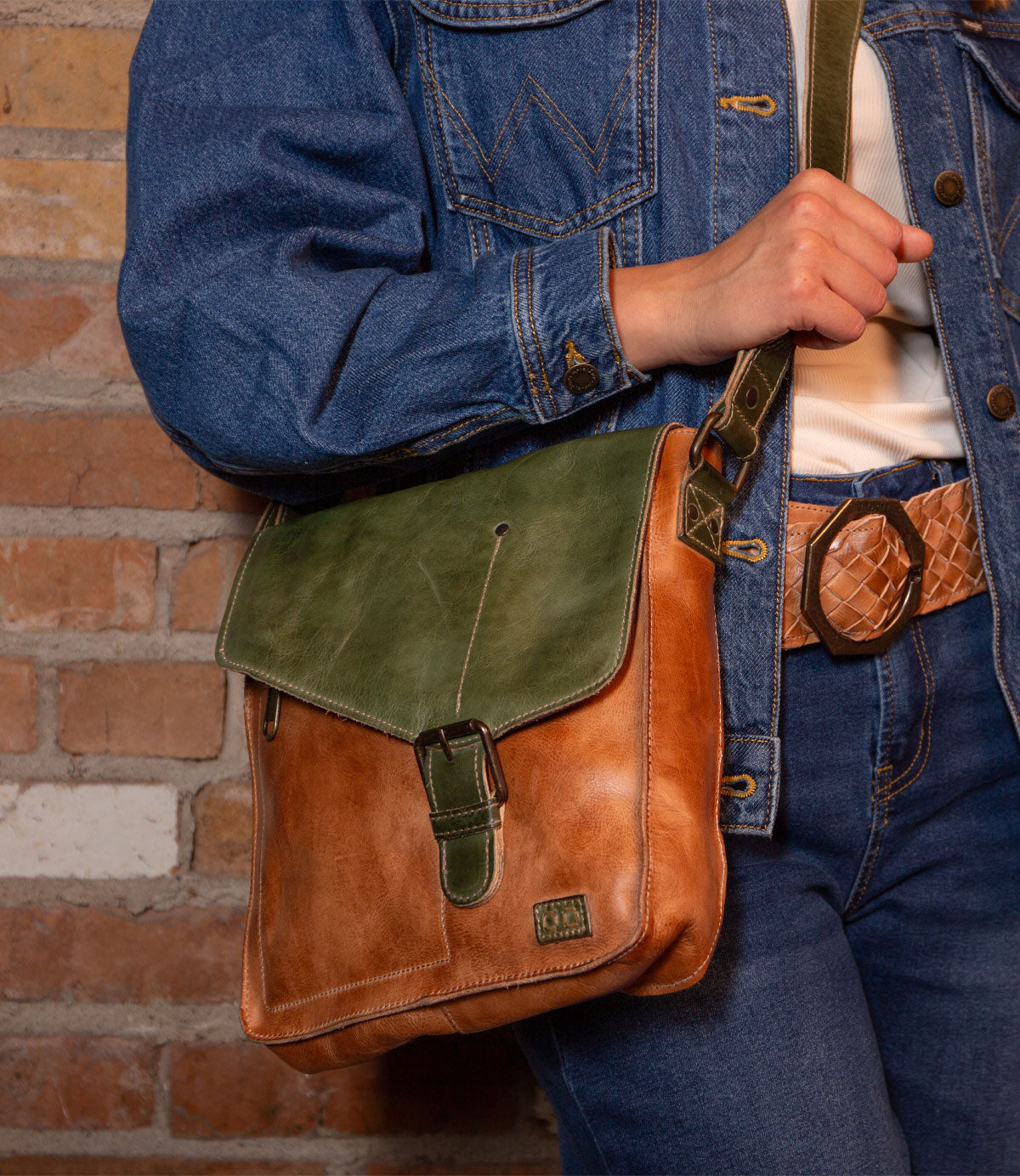 A person wearing a denim jacket and jeans stands against a brick wall, holding the Bed|Stü Venice Beach crossbody bag in brown and green, crafted from full-grain leather with an adjustable strap and buckle.