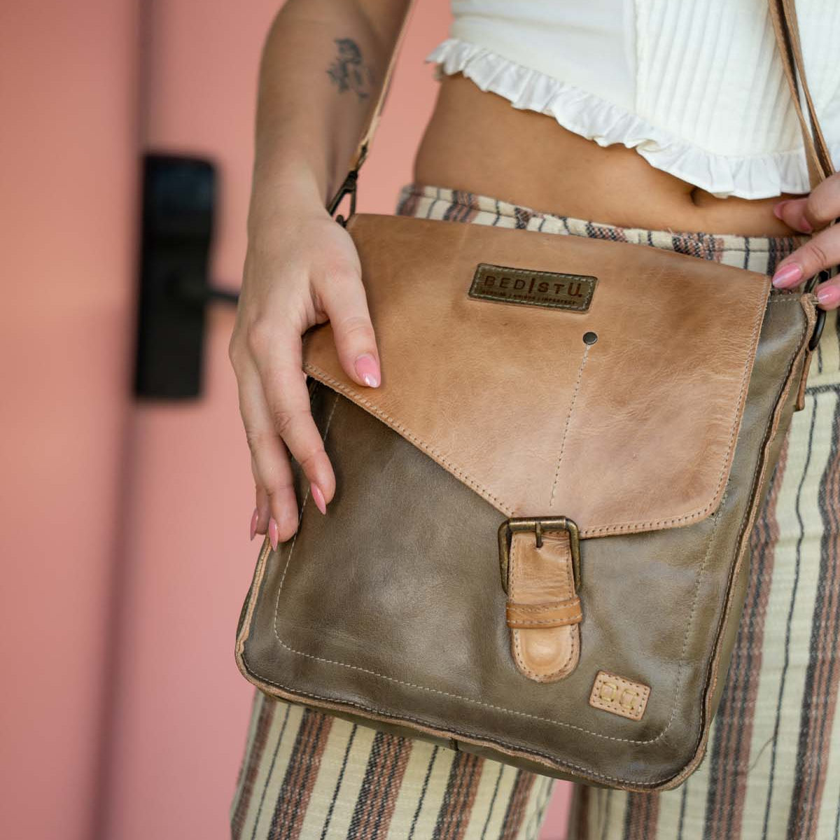 A person in striped pants and a white top holds the Bed|Stü Venice Beach crossbody bag, crafted from tan and olive full-grain leather with an adjustable strap, buckle detail, and a "Bed|Stü" logo patch on the front.