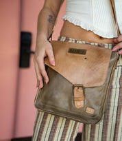 A person in striped pants and a white top holds the Bed|Stü Venice Beach crossbody bag, crafted from tan and olive full-grain leather with an adjustable strap, buckle detail, and a "Bed|Stü" logo patch on the front.