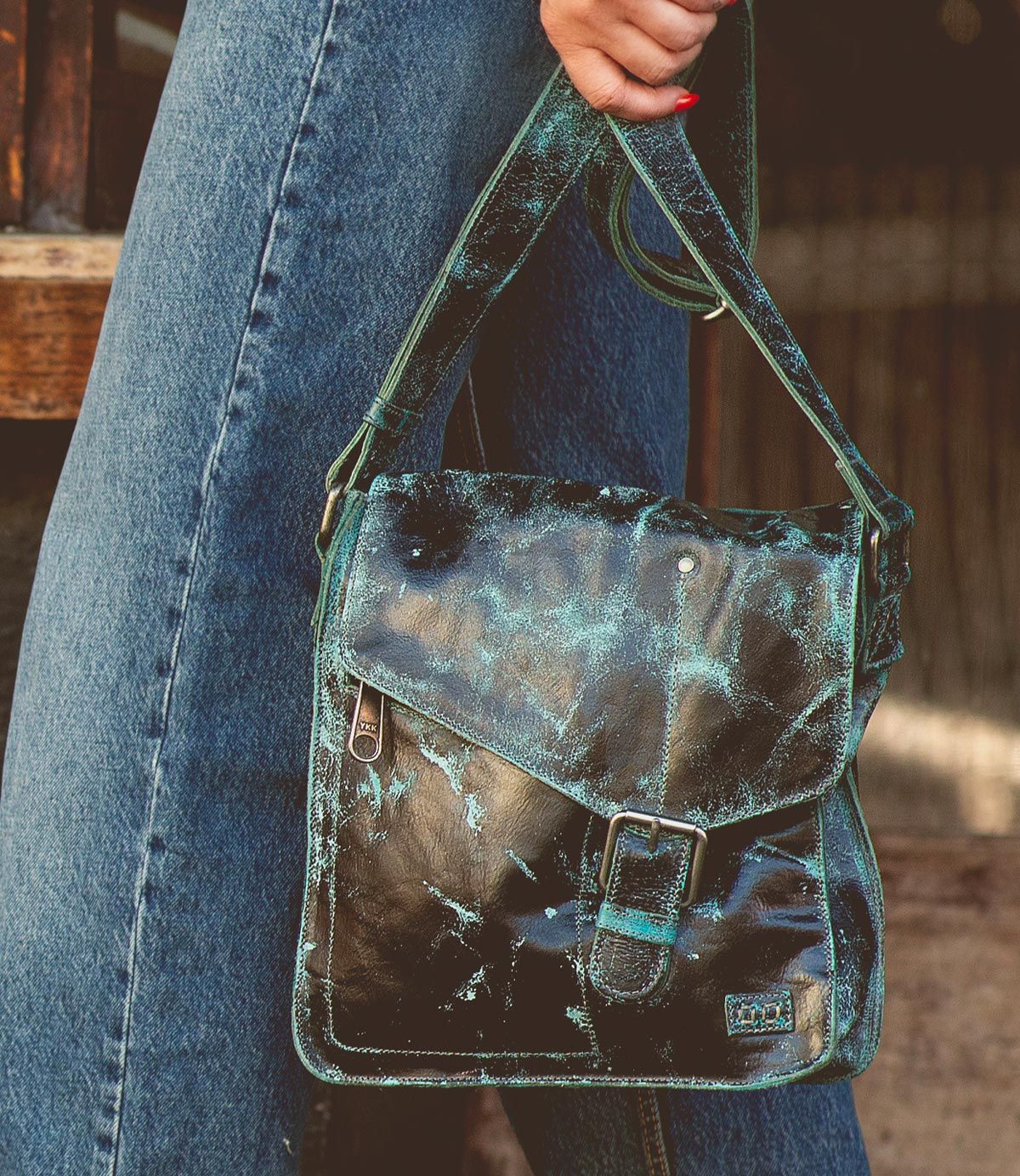 A person wearing jeans holds the Bed|Stü Venice Beach, a worn black full-grain leather crossbody bag featuring a blue-green distressed finish, front buckle detail, and an adjustable strap.