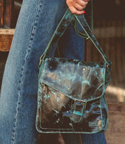 A person wearing jeans holds the Bed|Stü Venice Beach, a worn black full-grain leather crossbody bag featuring a blue-green distressed finish, front buckle detail, and an adjustable strap.