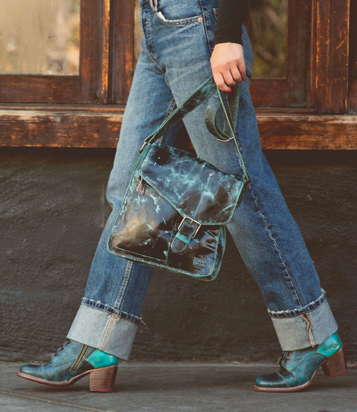 Wearing cuffed jeans and teal lace-up shoes, the person carries the Bed|Stü Venice Beach crossbody bag—a full-grain leather accessory in teal with a buckle and adjustable strap—as they walk past a dark wall and wooden window.