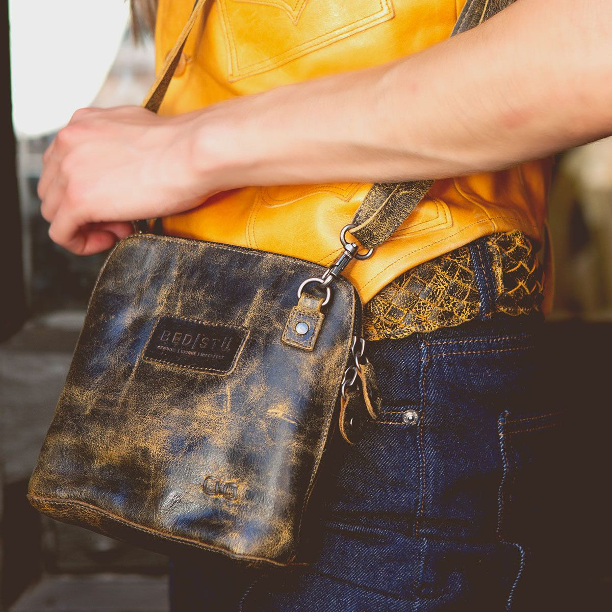 A person wearing a yellow shirt and blue jeans is holding the Ventura crossbody bag by Bed|Stü, which features a distressed leather finish, an exterior zip pocket, and a visible brand label.