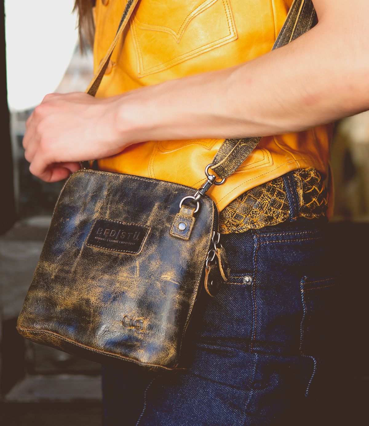 A person wearing a yellow shirt and blue jeans is holding the Ventura crossbody bag by Bed|Stü, which features a distressed leather finish, an exterior zip pocket, and a visible brand label.