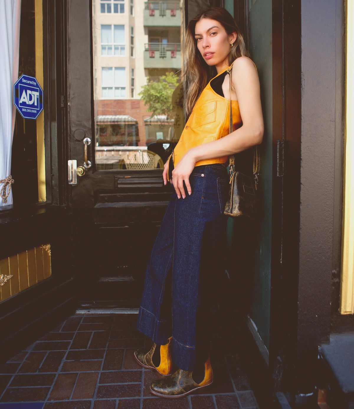 A person stands in a doorway on a city street wearing a yellow leather top, dark blue wide-leg jeans, and brown boots, holding the Bed|Stü Ventura crossbody bag featuring an exterior zip pocket.