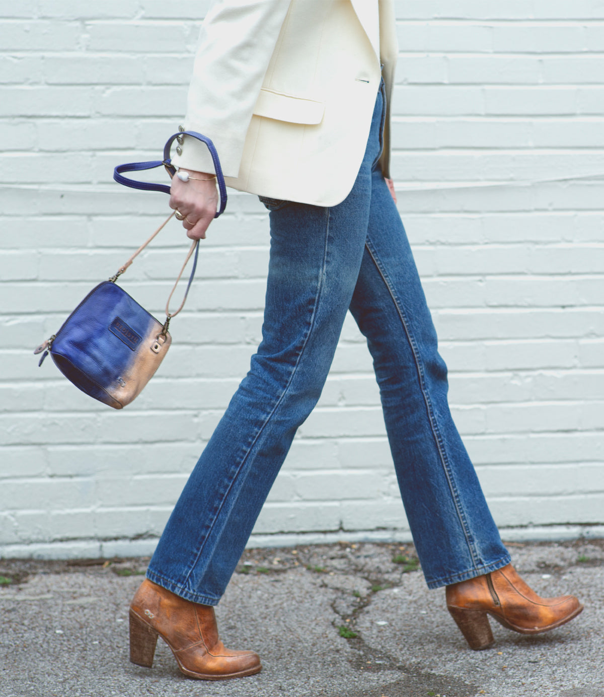 A person in blue jeans, tan heeled boots, and a white blazer walks on pavement, carrying the Bed|Stü Ventura lightweight purse with an exterior zip pocket in blue and gold by the strap.
