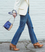 A person in blue jeans, tan heeled boots, and a white blazer walks on pavement, carrying the Bed|Stü Ventura lightweight purse with an exterior zip pocket in blue and gold by the strap.