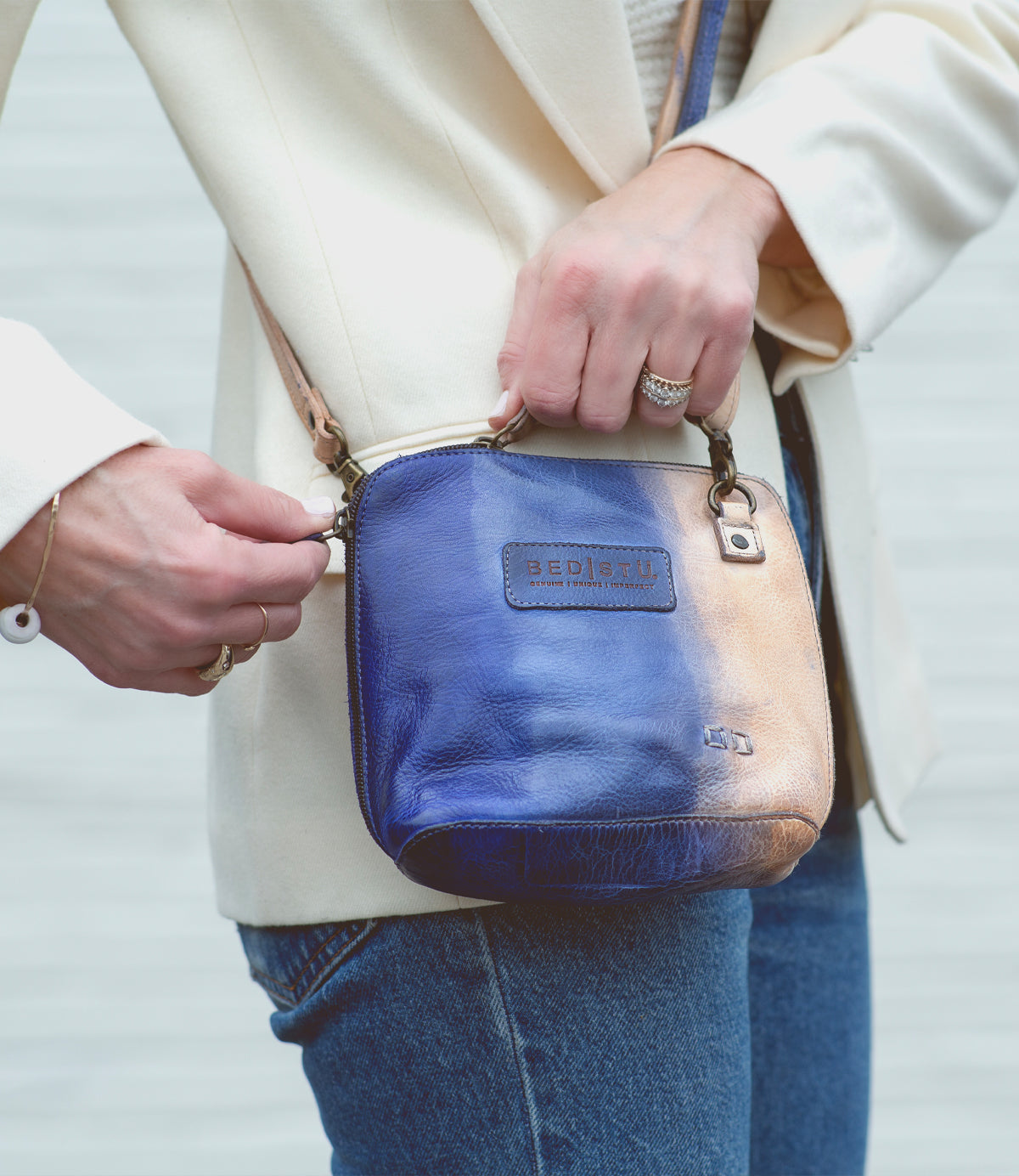 A person wearing a cream blazer and jeans holds the Bed|Stü Ventura, a small blue and tan leather crossbody bag featuring a metal zipper and clasp, as well as an exterior zip pocket for added convenience.