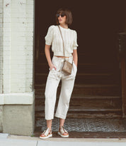A woman stands in front of a building entrance with her hands in her pockets, wearing sunglasses, a white knit top, cropped white pants, sandals, and the Bed|Stü Ventura leather crossbody bag featuring an exterior zip pocket.