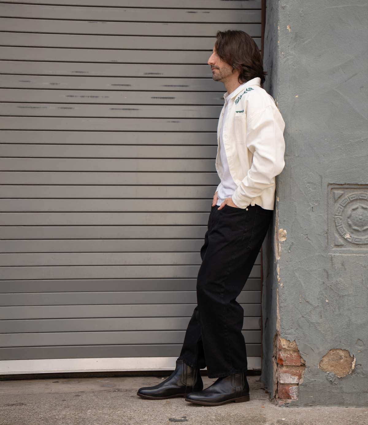 A man in a white jacket, black pants, and Bed|Stü Verrocchio handcrafted leather Chelsea boots leans against a gray wall next to a closed metal garage door, looking to the side.