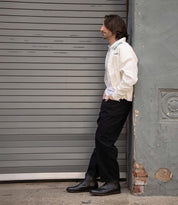A man in a white jacket, black pants, and Bed|Stü Verrocchio handcrafted leather Chelsea boots leans against a gray wall next to a closed metal garage door, looking to the side.
