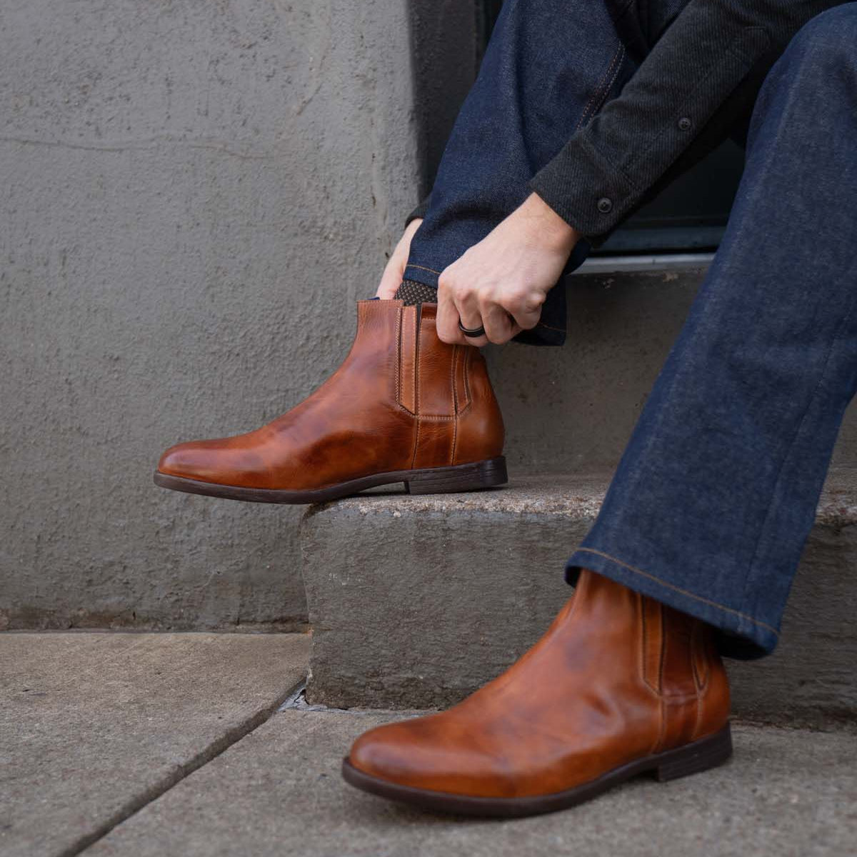 A person wearing blue jeans and brown Bed|Stü Verrocchio handcrafted leather Chelsea boots sits on concrete steps, adjusting one boot with their hand—perfect for everyday wear.