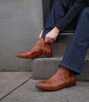 A person wearing blue jeans and brown Bed|Stü Verrocchio handcrafted leather Chelsea boots sits on concrete steps, adjusting one boot with their hand—perfect for everyday wear.