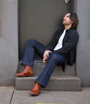 A man with dark hair and a beard sits on a concrete step in front of a door, dressed in everyday wear—a dark jacket, white shirt, blue jeans, and handcrafted Bed|Stü Verrocchio leather Chelsea boots. He looks up thoughtfully.