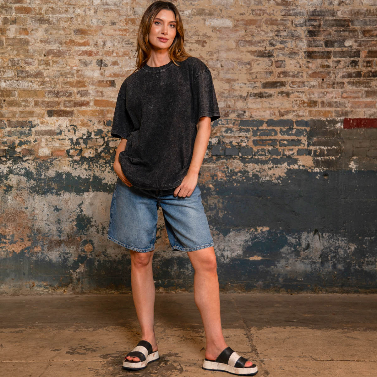 A person stands indoors against a textured brick wall, wearing a loose black t-shirt, denim shorts, and the Bed|Stü Vext stylish black-and-white platform slide sandals featuring a leather-wrapped sole.