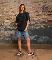 A person stands indoors against a textured brick wall, wearing a loose black t-shirt, denim shorts, and the Bed|Stü Vext stylish black-and-white platform slide sandals featuring a leather-wrapped sole.