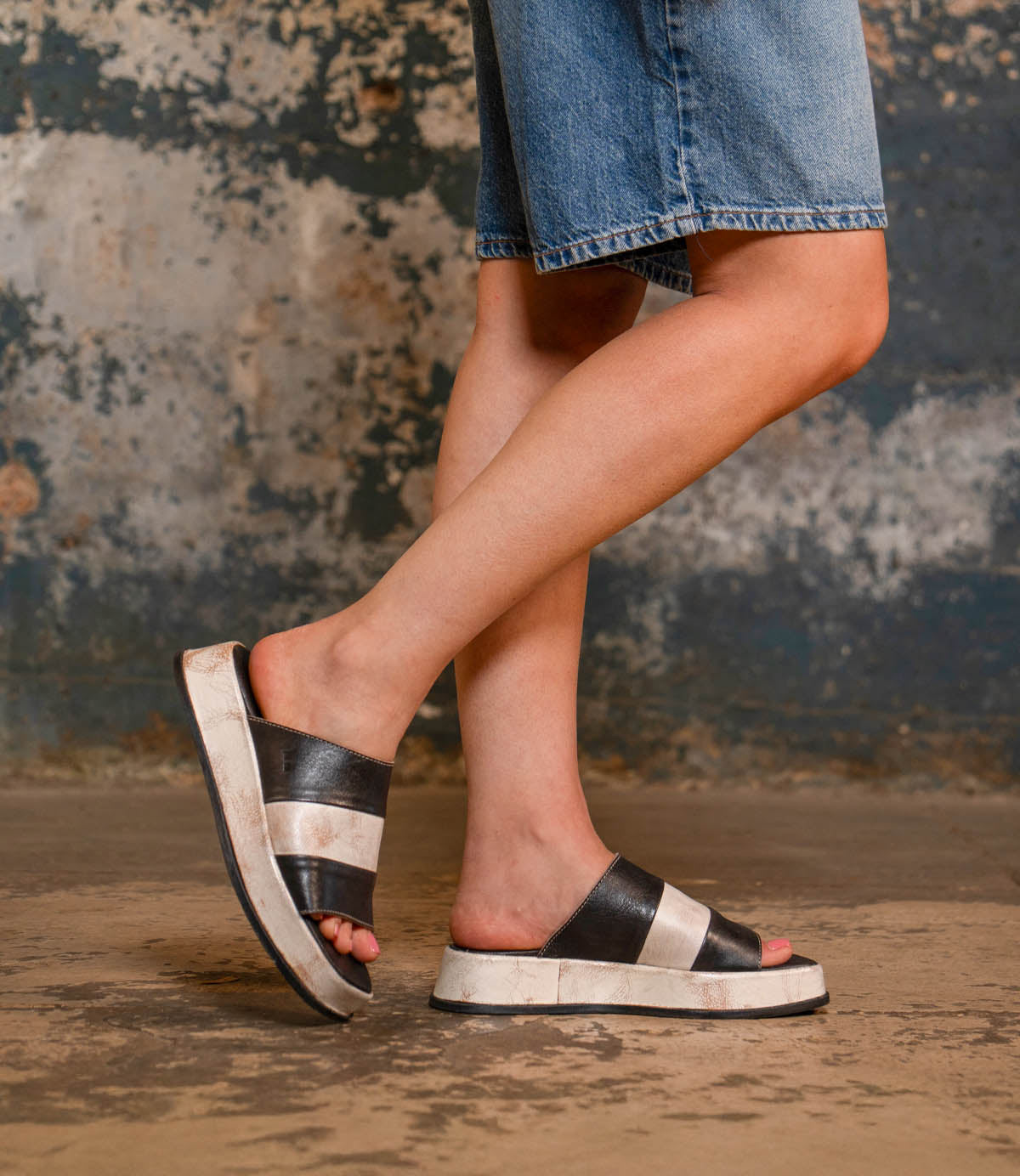 A person wearing the Bed|Stü Vext black and white square-toe sandals and denim shorts stands on a concrete floor against a textured wall.