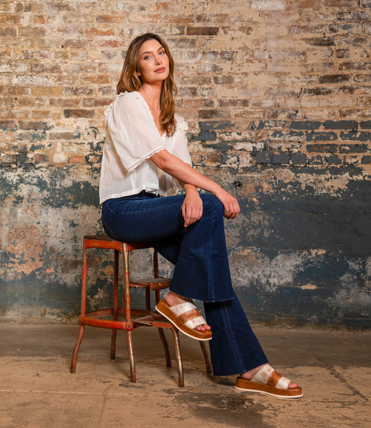 A woman wearing the Bed|Stü Vext brown platform slide sandals, featuring a leather-wrapped sole, sits on a stool against a worn brick wall in a white blouse and blue jeans, looking at the camera.